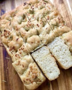 Sliced Italian focaccia bread on a wooden chopping board, showcasing its crispy crust and soft, airy interior, drizzled with olive oil and sprinkled with rosemary and sea salt