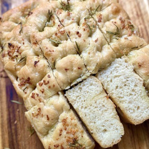 Sliced Italian focaccia bread on a wooden chopping board, showcasing its crispy crust and soft, airy interior, drizzled with olive oil and sprinkled with rosemary and sea salt