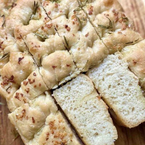 Sliced Italian focaccia bread on a wooden chopping board, showcasing its crispy crust and soft, airy interior, drizzled with olive oil and sprinkled with rosemary and sea salt