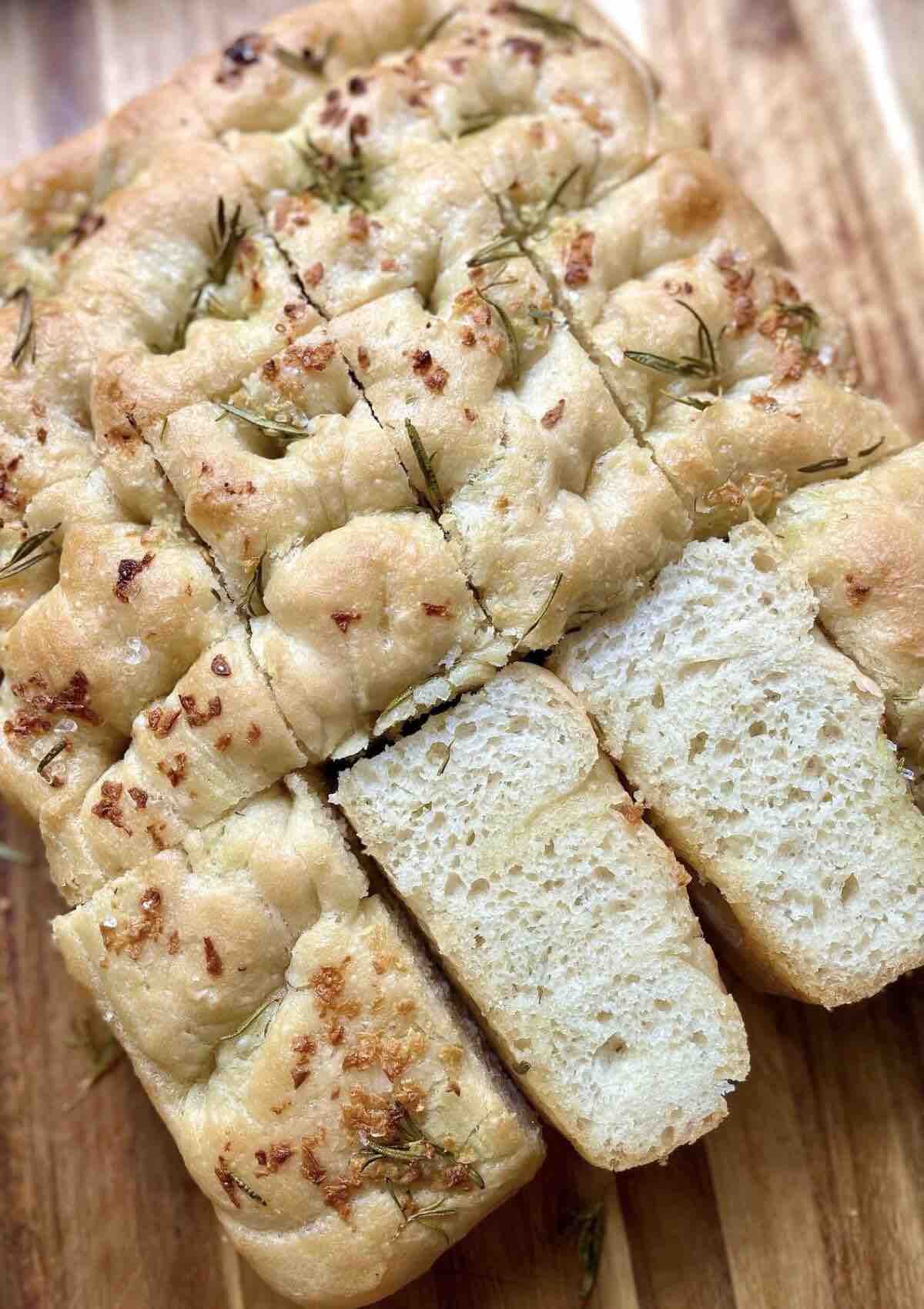 Sliced Italian focaccia bread on a wooden chopping board, showcasing its crispy crust and soft, airy interior, drizzled with olive oil and sprinkled with rosemary and sea salt