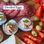 An appetizing plate with deviled eggs, salami slices, green olives, sliced tomatoes, cheese triangles, and a slice of bread with a generous egg filling spread.