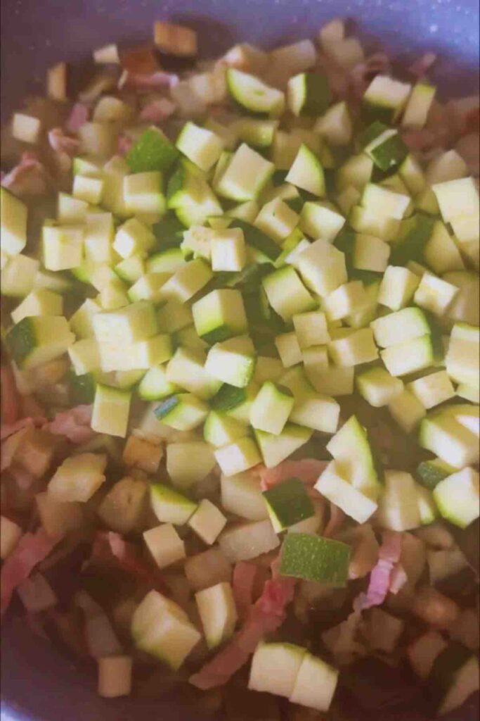 Olive oil in a pan with cubed potatoes, eggplant, bacon strips, and cubed zucchini, all sautéing together.