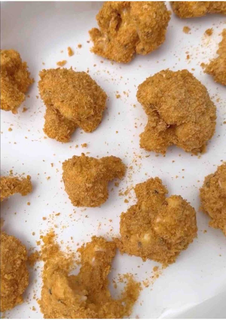 Cauliflower bites coated in batter and crushed cornflakes spread out in a pan, ready to be baked.