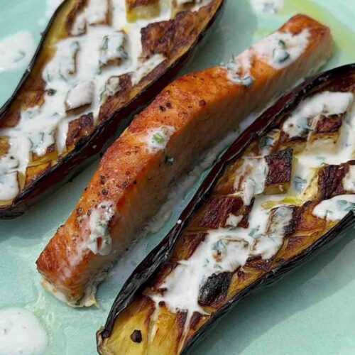 Two halves of baked aubergine with yogurt dressing served on a plate alongside roasted salmon