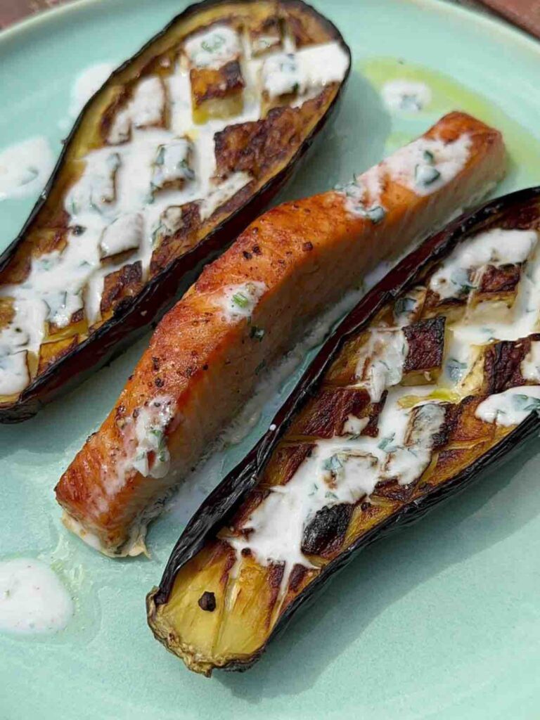 Two halves of baked aubergine with yogurt dressing served on a plate alongside roasted salmon