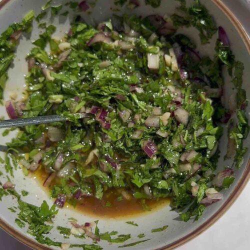 Chimichurri sauce in a bowl, featuring finely chopped red onion, fresh parsley, fresh coriander, grated garlic, red wine vinegar, olive oil, diced chili pepper, and seasoned with salt and pepper.