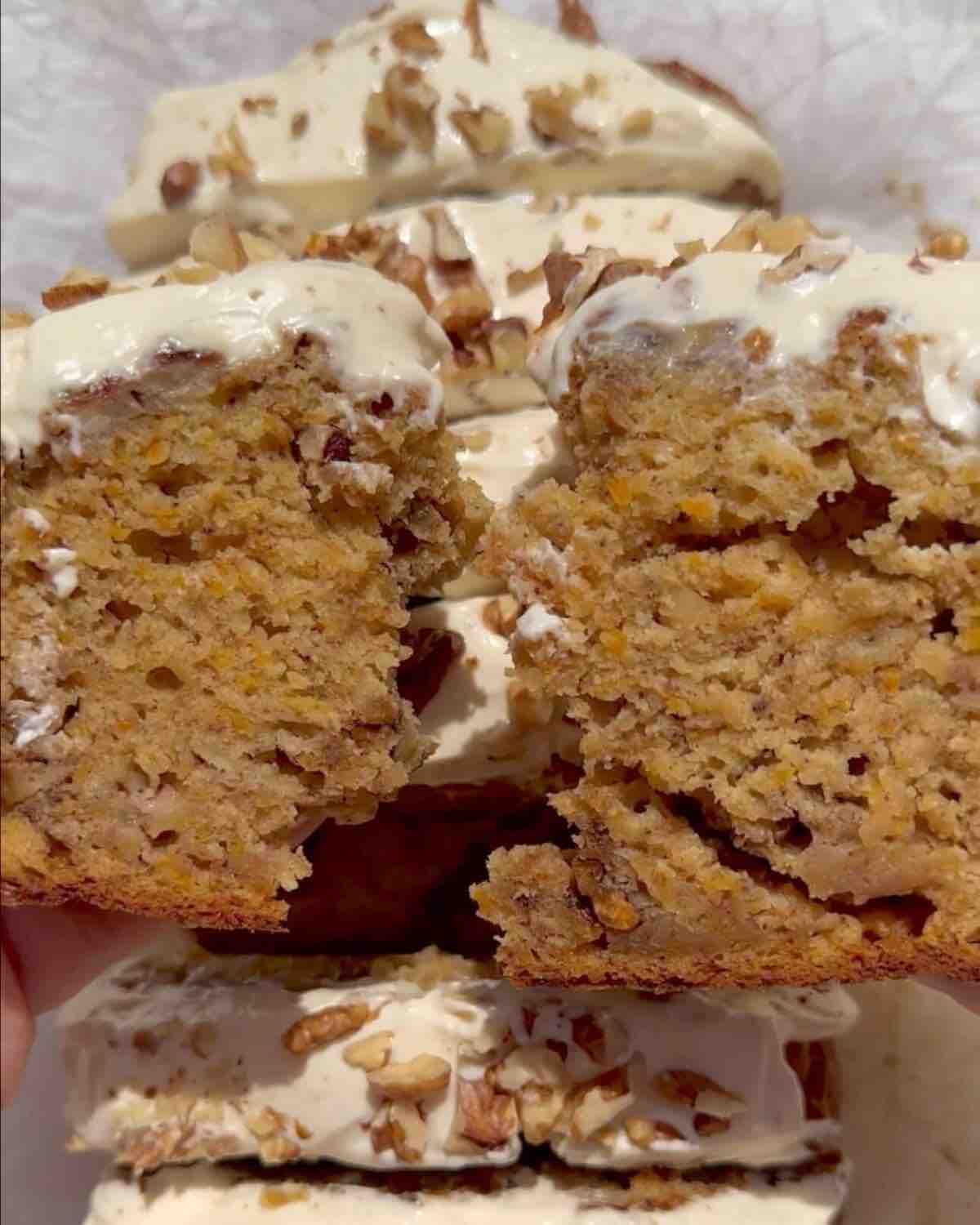 A vegan carrot cake banana bread topped with cream cheese frosting and walnuts. In the foreground, a slice of the bread is broken in two, revealing its moist texture, while the background shows the full banana loaf, neatly sliced.