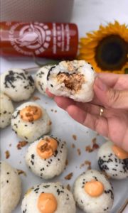 In the foreground, a plate displays a rice ball generously sprinkled with sesame seeds, revealing a tantalizing tuna filling. Adjacent to it, a plate of tuna onigiri adds to the culinary array. In the backdrop, a bottle of sriracha sauce stands beside a vibrant sunflower, completing the picturesque scene.