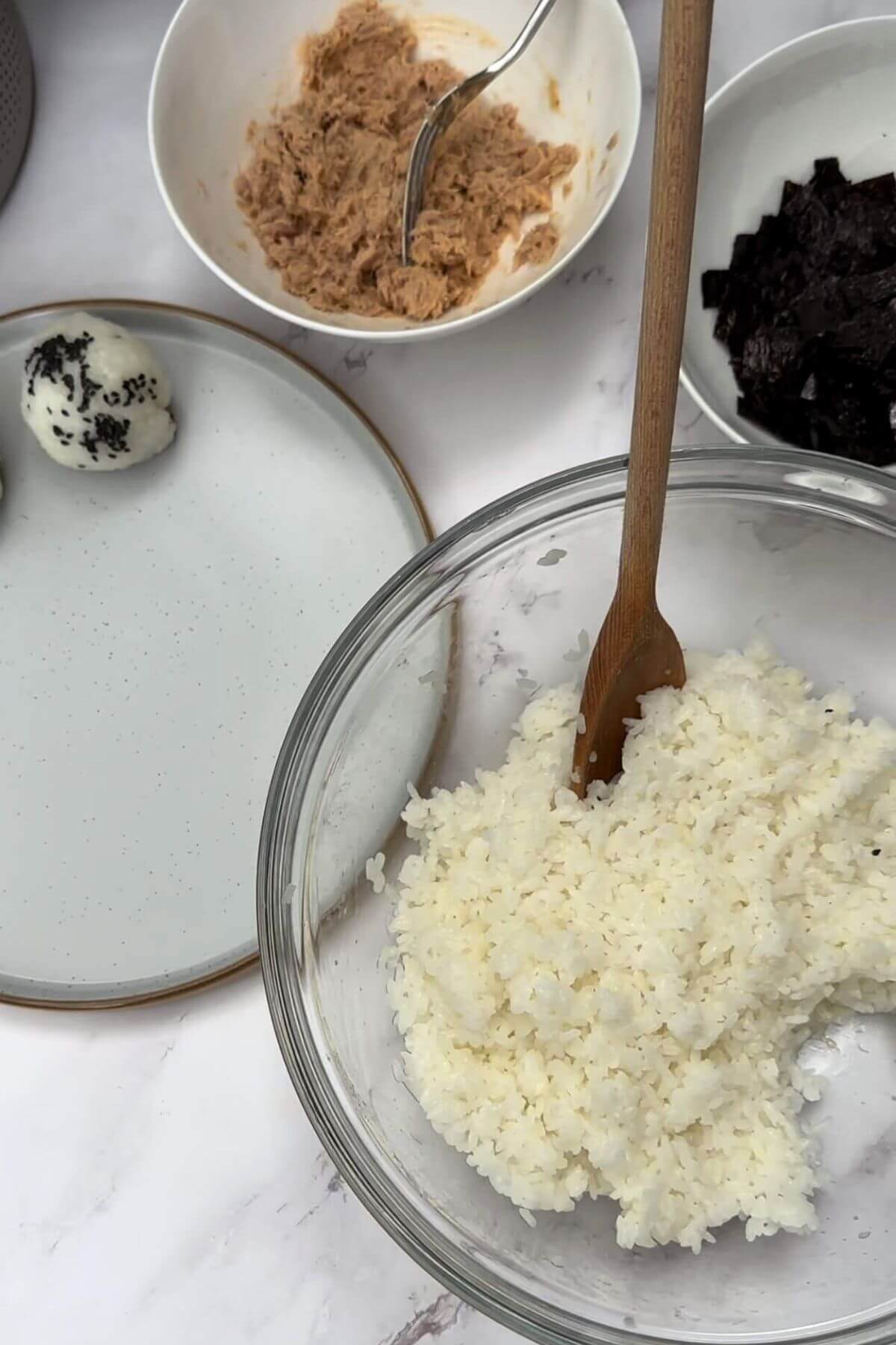 A picture of food on a table. There's a bowl of tuna, a bowl of rice, a bowl of seaweed cut into small pieces, and a tuna onigiri on a plate.