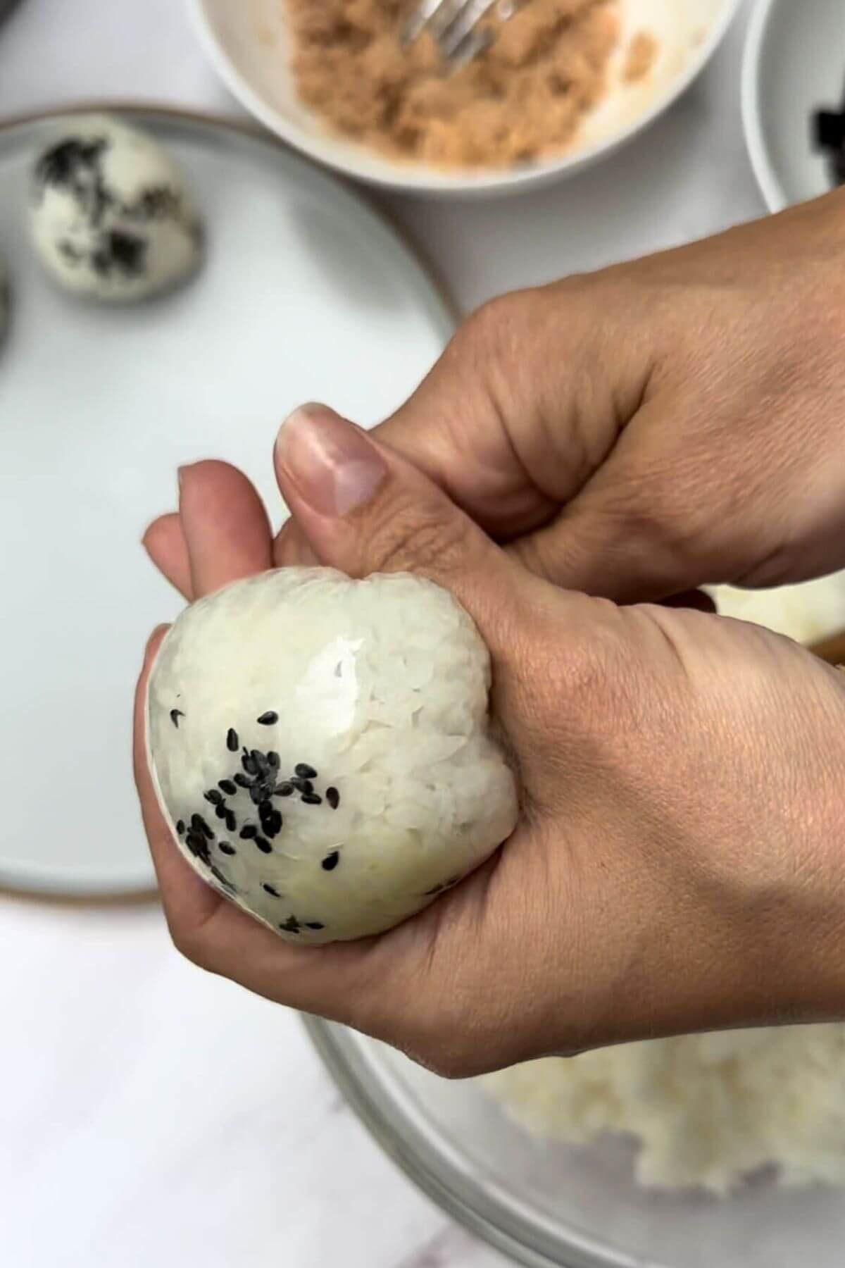 In the picture, you can see a table with different bowls and plates. There's a bowl of tuna, a bowl of rice, and a bowl of seaweed cut into small strips. On a plate, there's a special rice ball called onigiri with tuna inside. In the middle of the table, there's a hand holding a round-shaped onigiri wrapped in plastic wrap to make it look like a ball.