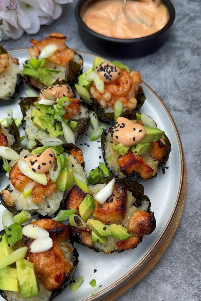 A picture of a plate on a table with baked salmon and prawn sushi cups. The dish is topped with green onions, avocado pieces, spicy mayo, and sesame seeds.