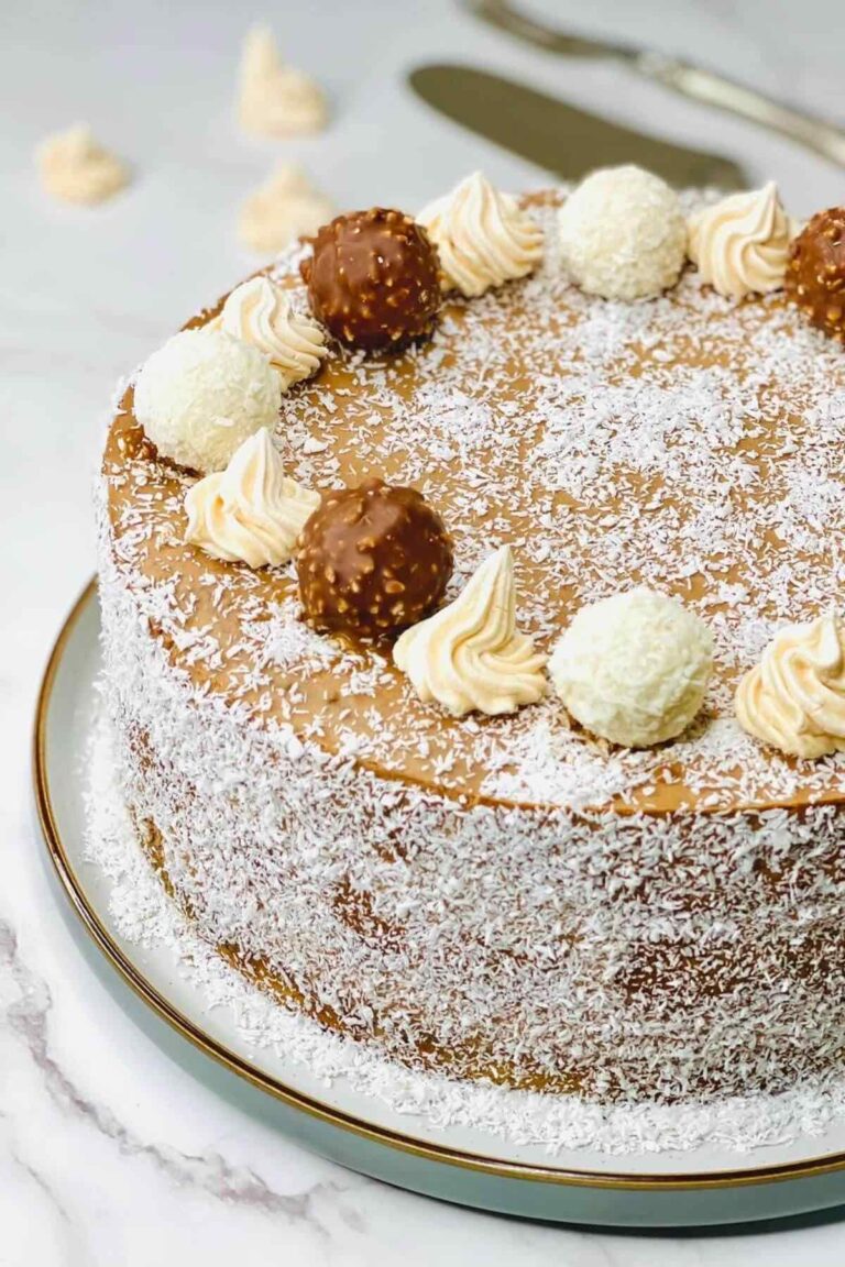 A close-up of a rich dark chocolate orange cake topped with smooth chocolate cream, sprinkled with coconut flakes, and decorated with Ferrero Rocher and Raffaello chocolates placed in a circle around the edge of the cake.