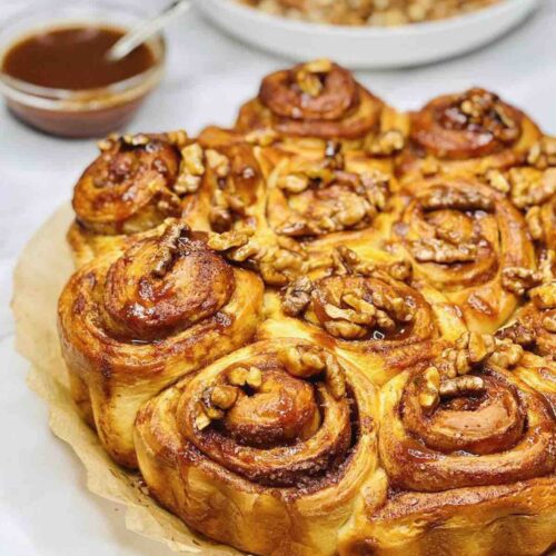 Biscoff Cinnamon Rolls with caramel sauce and walnuts in separate bowls in the background.