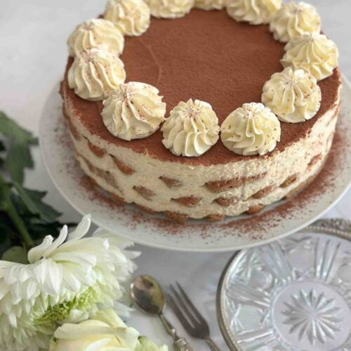 Tiramisu cake displayed on a white ceramic cake stand, with a white rose in front of the stand for decoration. The setting includes silver cutlery and an artisanal plate with silver trim.