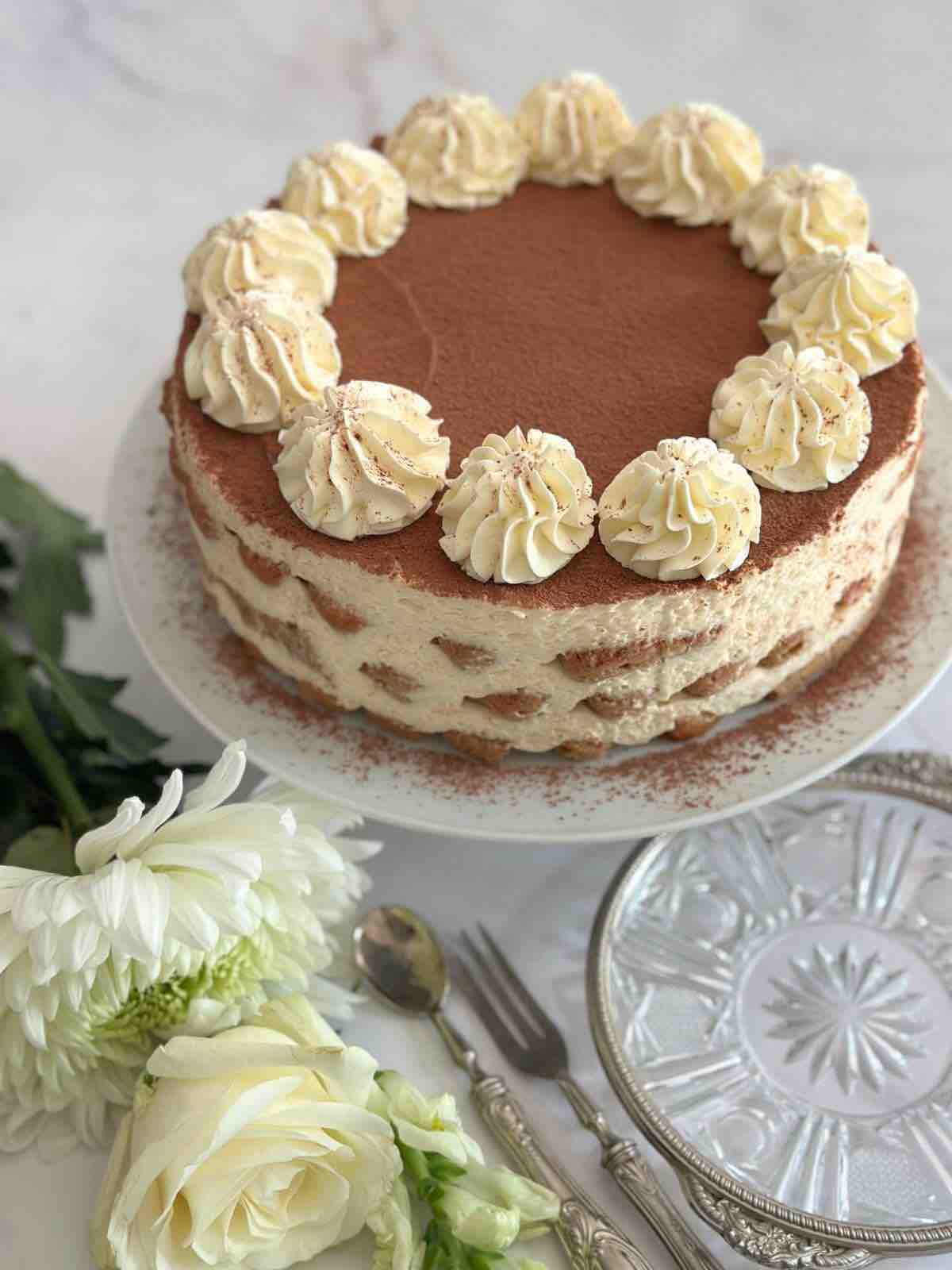 Tiramisu cake displayed on a white ceramic cake stand, with a white rose in front of the stand for decoration. The setting includes silver cutlery and an artisanal plate with silver trim.