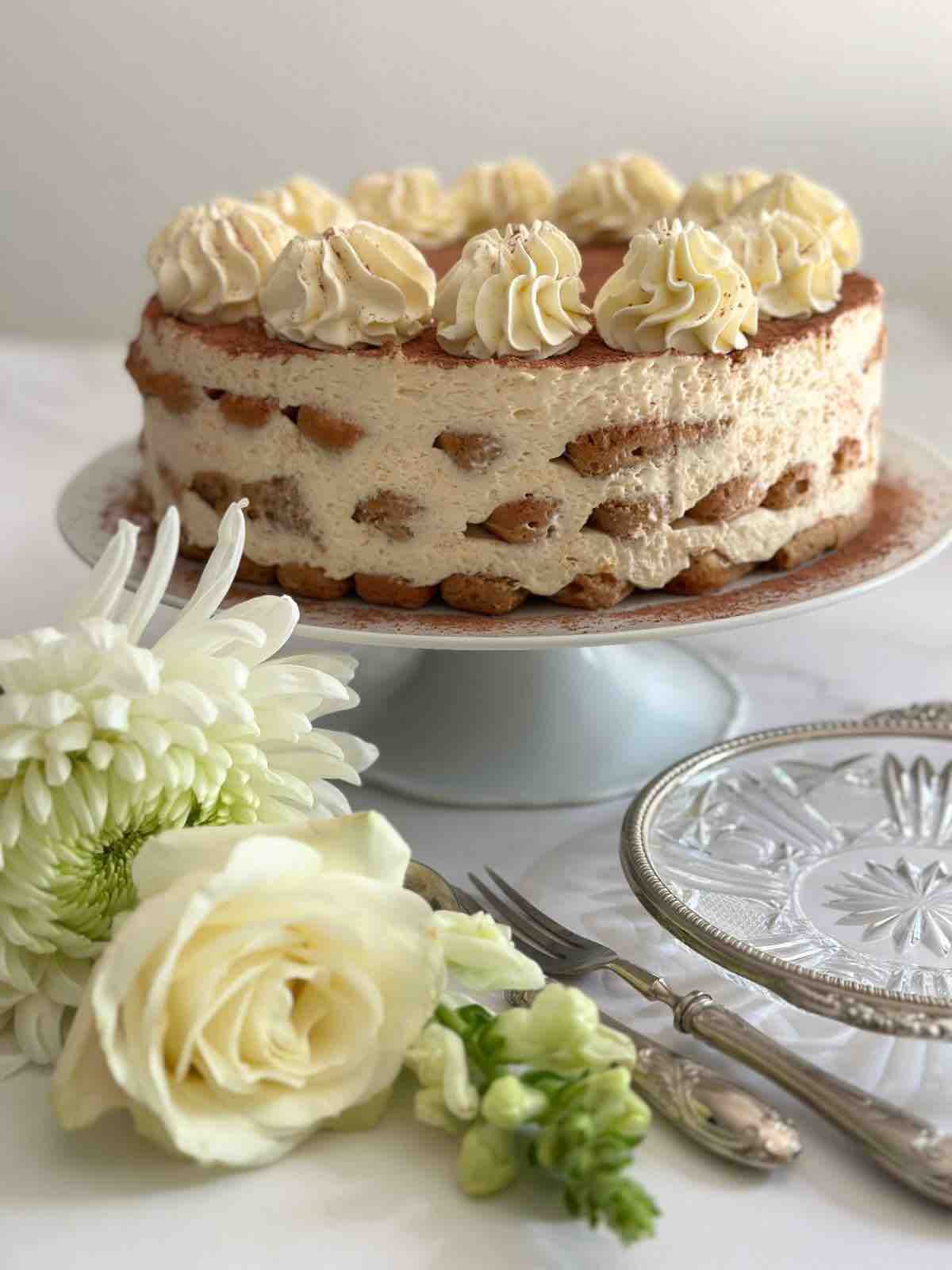 Tiramisu cake displayed on a white ceramic cake stand, with a white rose in front of the stand for decoration. The setting includes silver cutlery and an artisanal plate with silver trim.