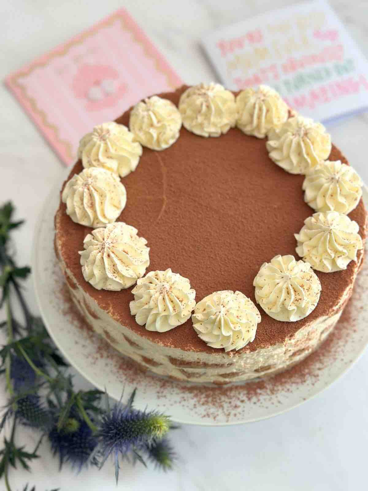 Tiramisu cake displayed on a white ceramic cake stand, with a blue flower in front of the stand for decoration. The setting includes 2 birthday cards in the background.