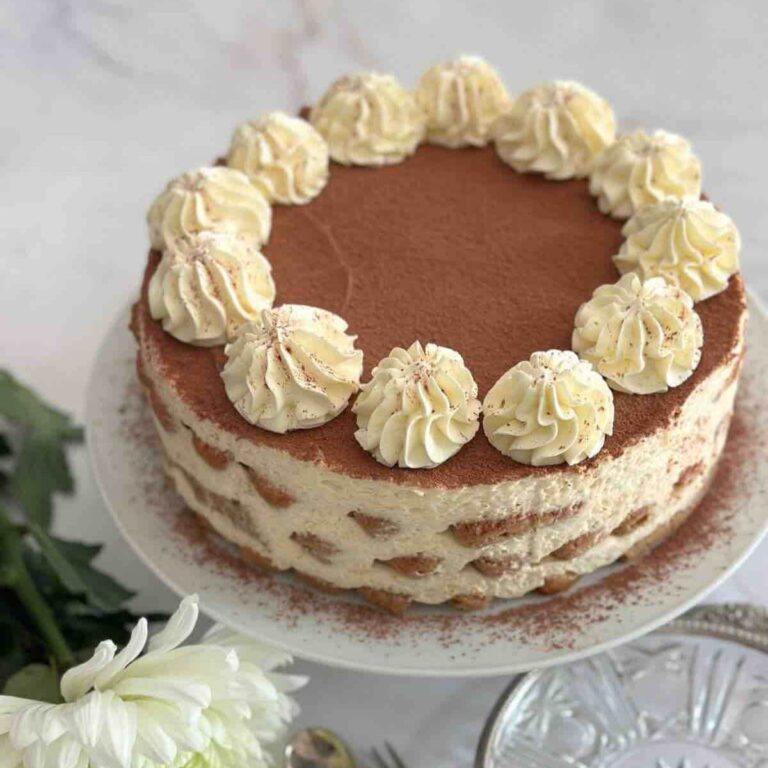 Tiramisu cake on a white stand, decorated with a white rose, silverware, and plate.