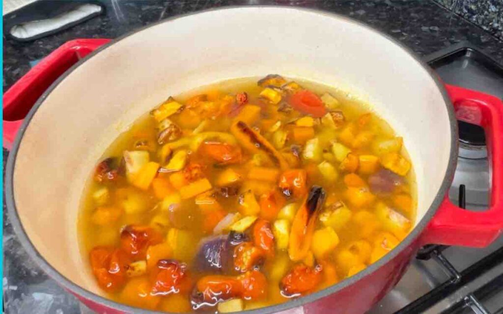 Roasted vegetables and garlic in a pot with broth and basil, cooking on the stove.