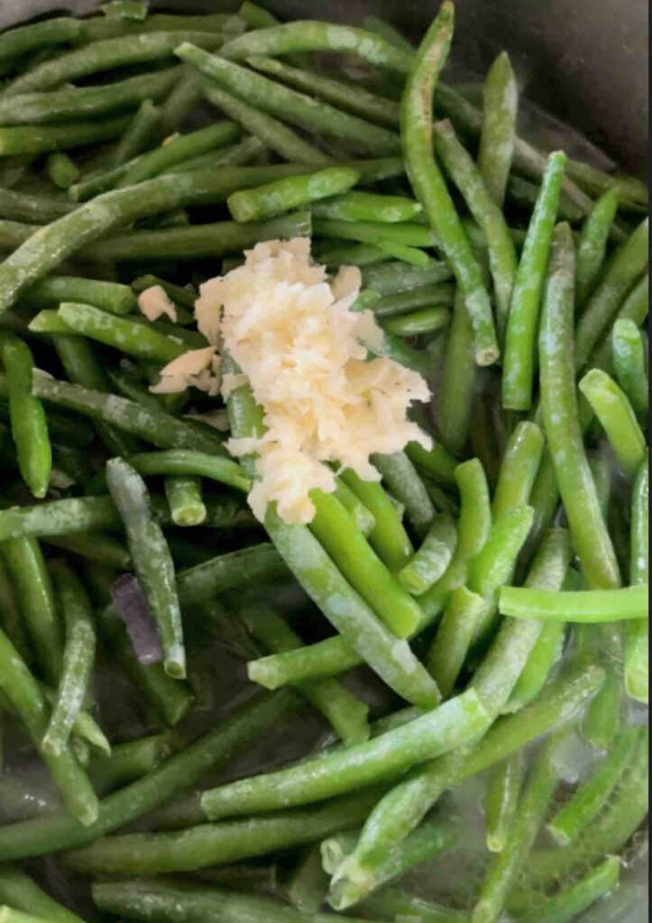 Onions, salt, pepper, and a stock cube sautéing in a pan with frozen green beans and crushed garlic added on top