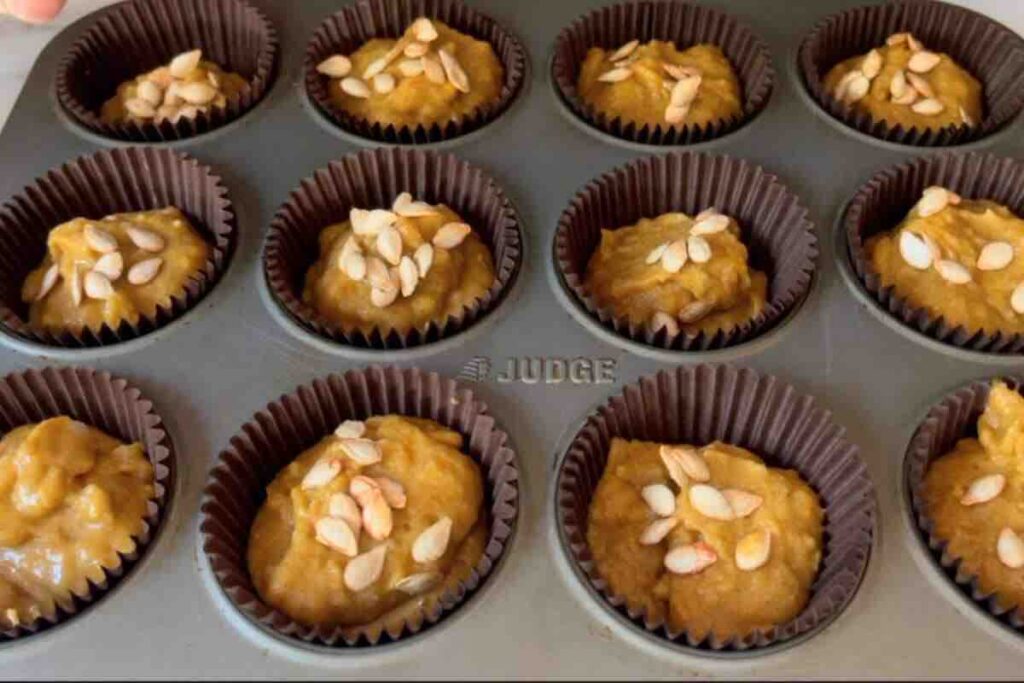 Batter evenly distributed in 12 cupcake liners, ready for baking, with pumpkin seeds sprinkled on top. 