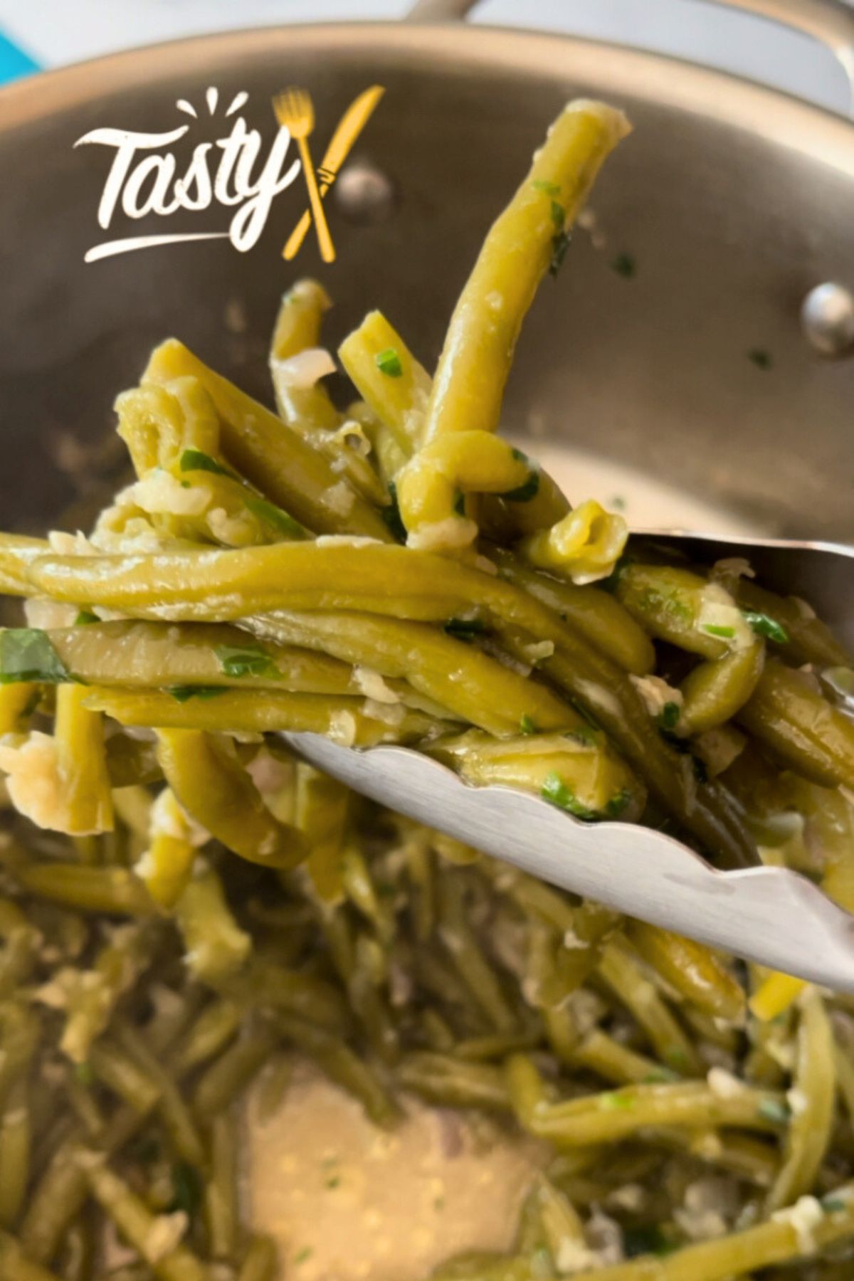 Sautéed green beans with garlic, garnished with chopped parsley, being lifted with kitchen tongs.