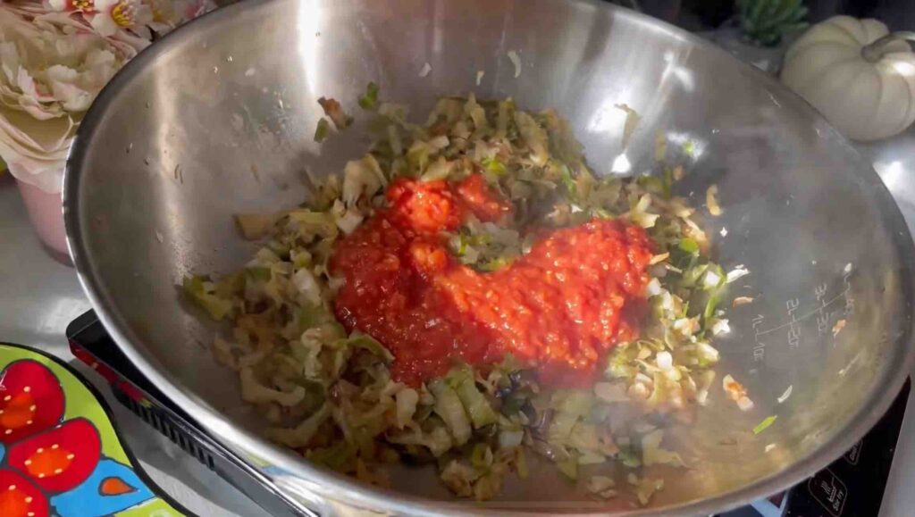 Chopped red onion sautéing in a wok with olive oil, with shredded cabbage added and tomato sauce being poured on top.