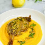 Roasted duck leg served with butternut squash puree on a white plate, garnished with lemon sauce and fresh green mint leaves.