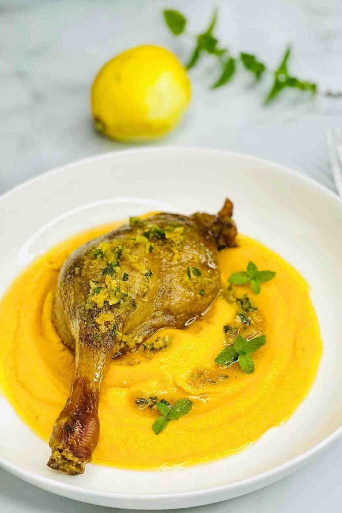 Roasted duck leg served with butternut squash puree on a white plate, garnished with lemon sauce and fresh green mint leaves.