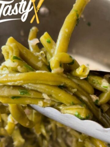 Sautéed green beans with garlic, garnished with chopped parsley, being lifted with kitchen tongs.