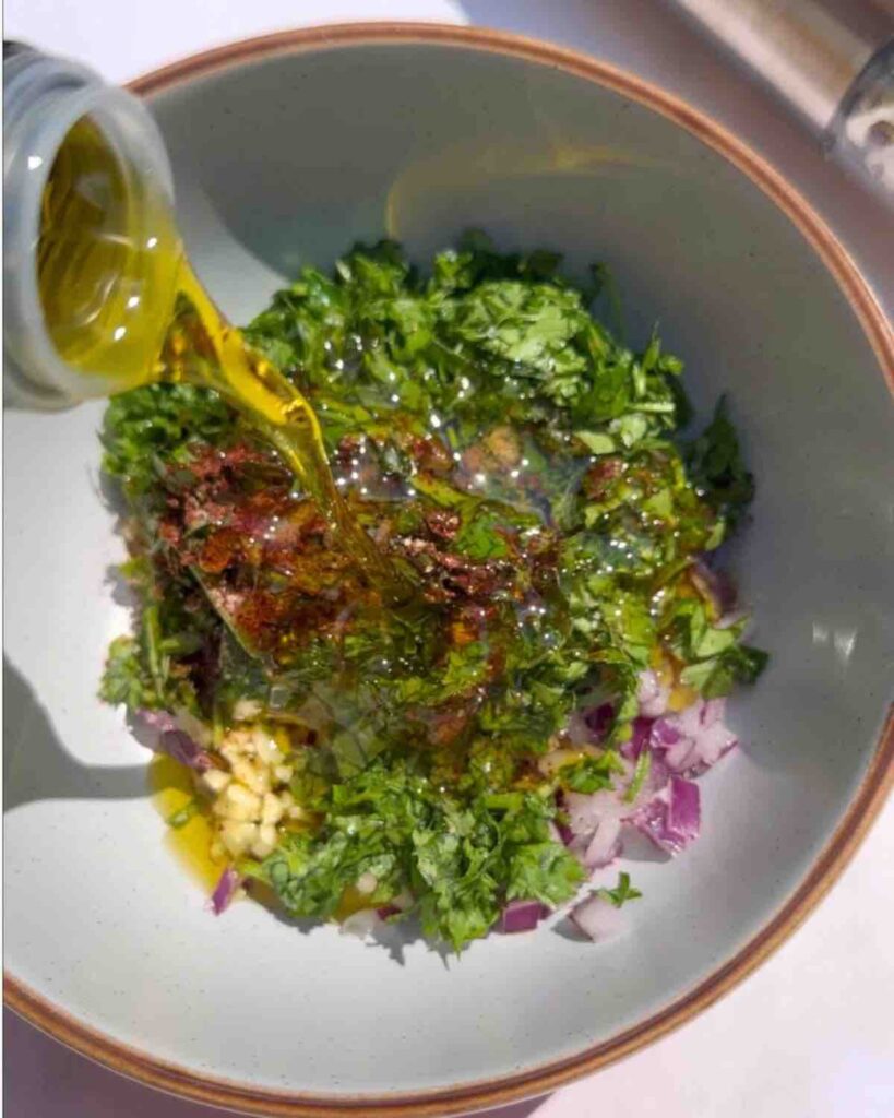 A bowl filled with chopped parsley, coriander, onion, garlic, chili pepper, and red wine vinegar. Olive oil is being poured into the bowl.