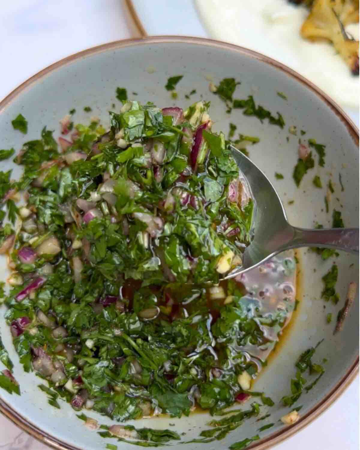 Chimichurri sauce in a bowl, made with finely chopped red onion, fresh parsley, coriander, grated garlic, red wine vinegar, olive oil, diced chili pepper, and seasoned with salt and pepper. A spoon full of the sauce is lifted above the bowl.