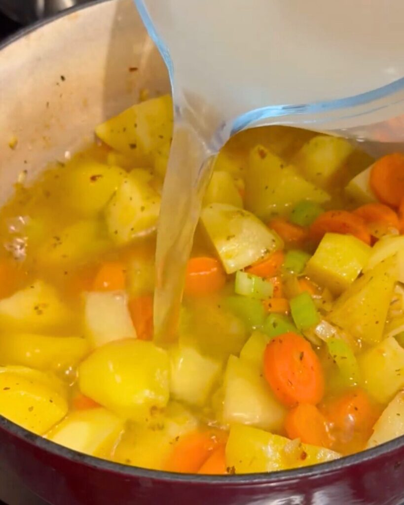 A deep pot with sautéed diced potatoes, carrots, onions, and sliced celery sticks, as chicken soup is being poured in to bring it to a boil.