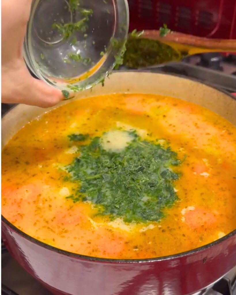 A deep pot filled with sautéed diced potatoes, carrots, onions, sliced celery sticks, and rice, simmering in chicken soup. The "Terbive" egg mix of plain flour, natural yogurt, egg yolk, and lemon juice is already stirred in, with parsley being added as garnish.