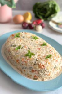 An oval plate filled with Olivier Salad (Boeuf Salad), garnished with mint leaves, set against a backdrop of fresh veggies.