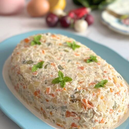 An oval plate filled with Olivier Salad (Boeuf Salad), garnished with mint leaves, set against a backdrop of fresh veggies.