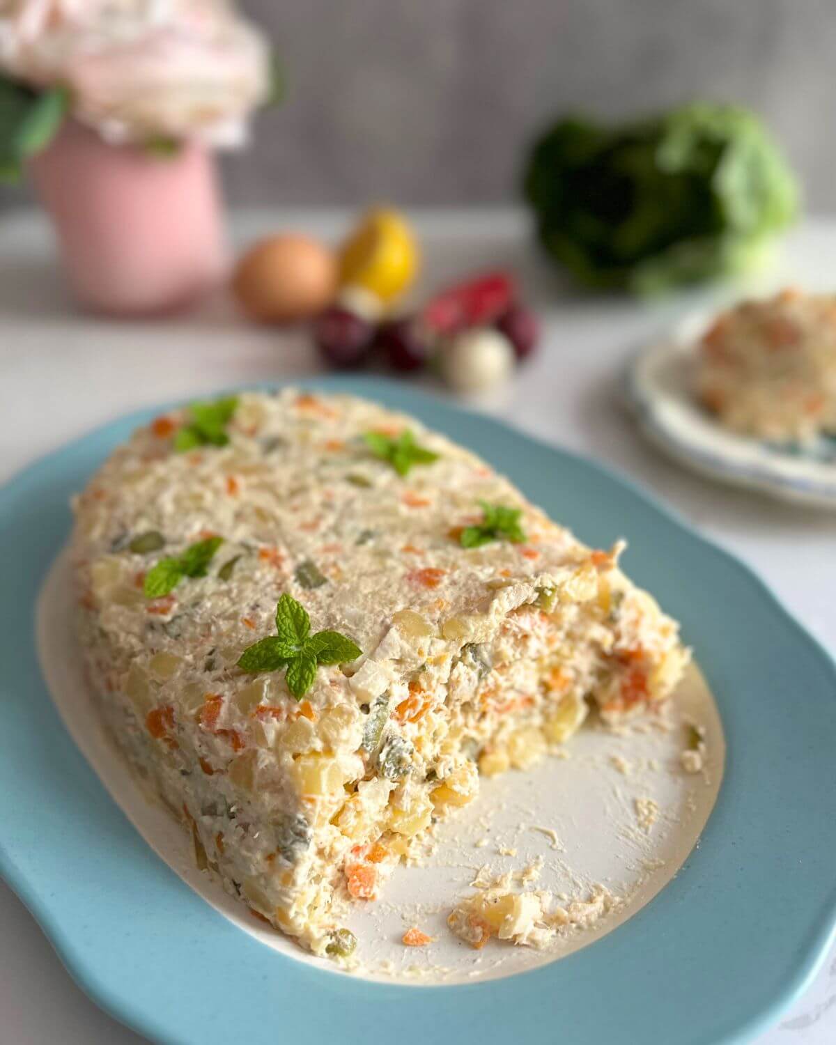 An oval plate filled with Olivier Salad (Boeuf Salad), garnished with mint leaves, is set against a backdrop of fresh veggies, with a piece taken out to reveal the center. 