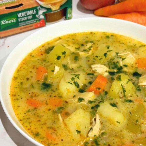 A delicious bowl of chicken soup garnished with herbs, served in a white shallow bowl, with colorful vegetables in the background.