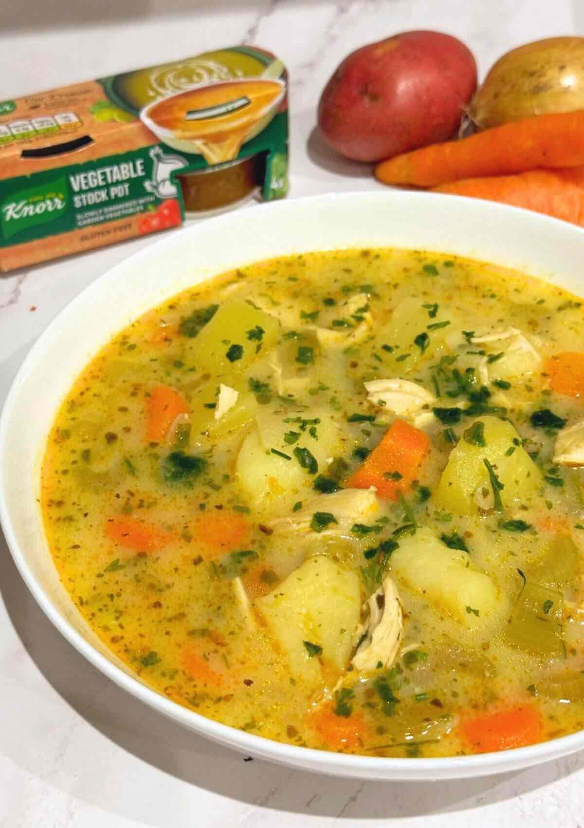 A delicious bowl of chicken soup garnished with herbs, served in a white shallow bowl, with colorful vegetables in the background.