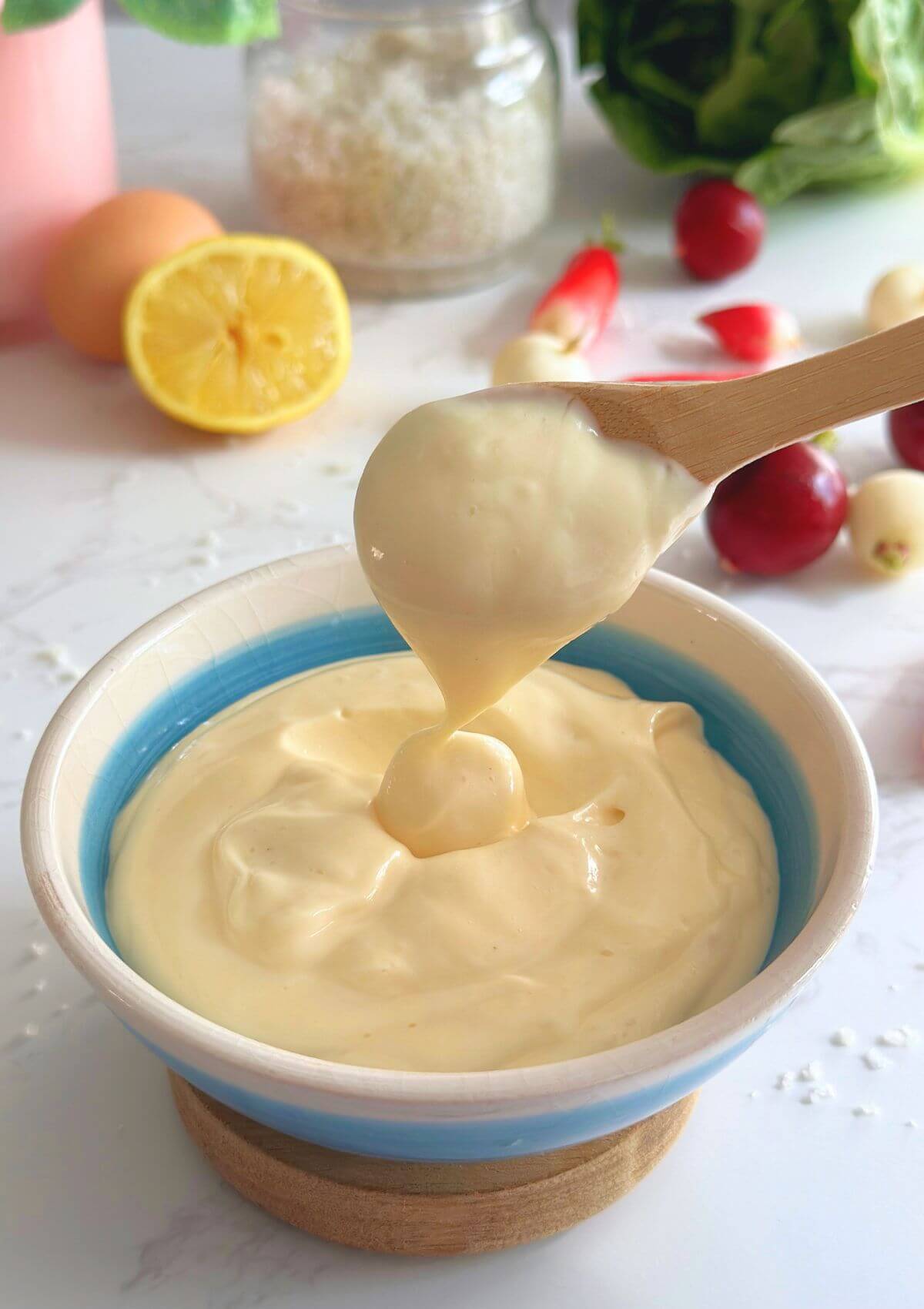 A bowl of homemade mayonnaise made with 4 ingredients, with a wooden spoon scooping up the creamy mayo to show its texture.