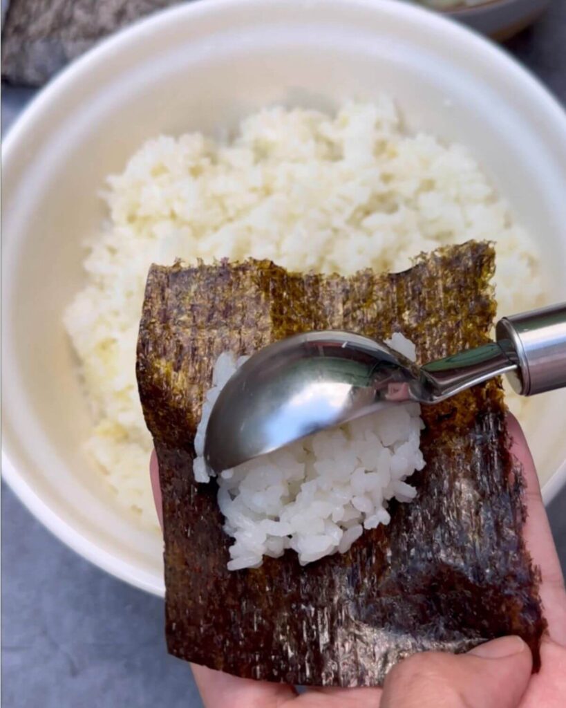 A close-up image showing a hand using an ice cream scoop to place a portion of seasoned sushi rice onto a square of nori seaweed, preparing to form sushi cups.