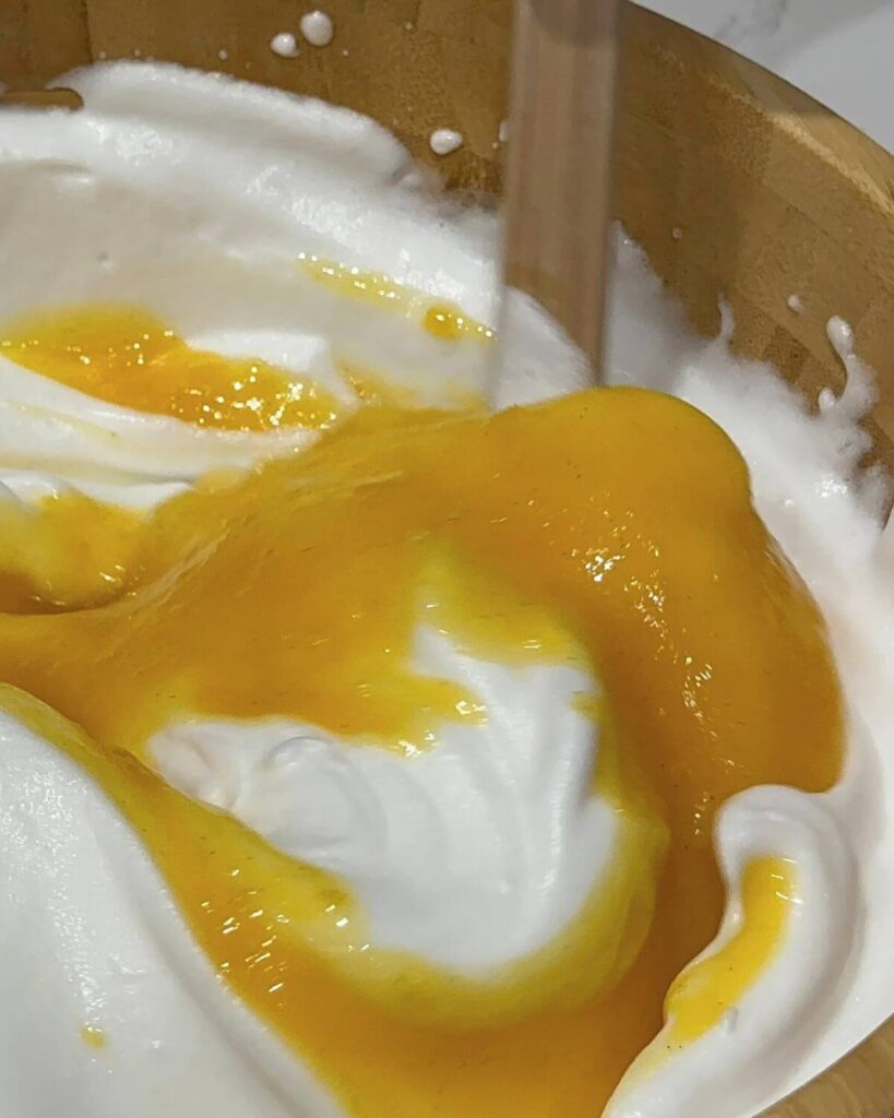 Gradually folding the yolk mixture into beaten egg whites using a balloon whisk, with a light and airy batter shown in a large mixing bowl.