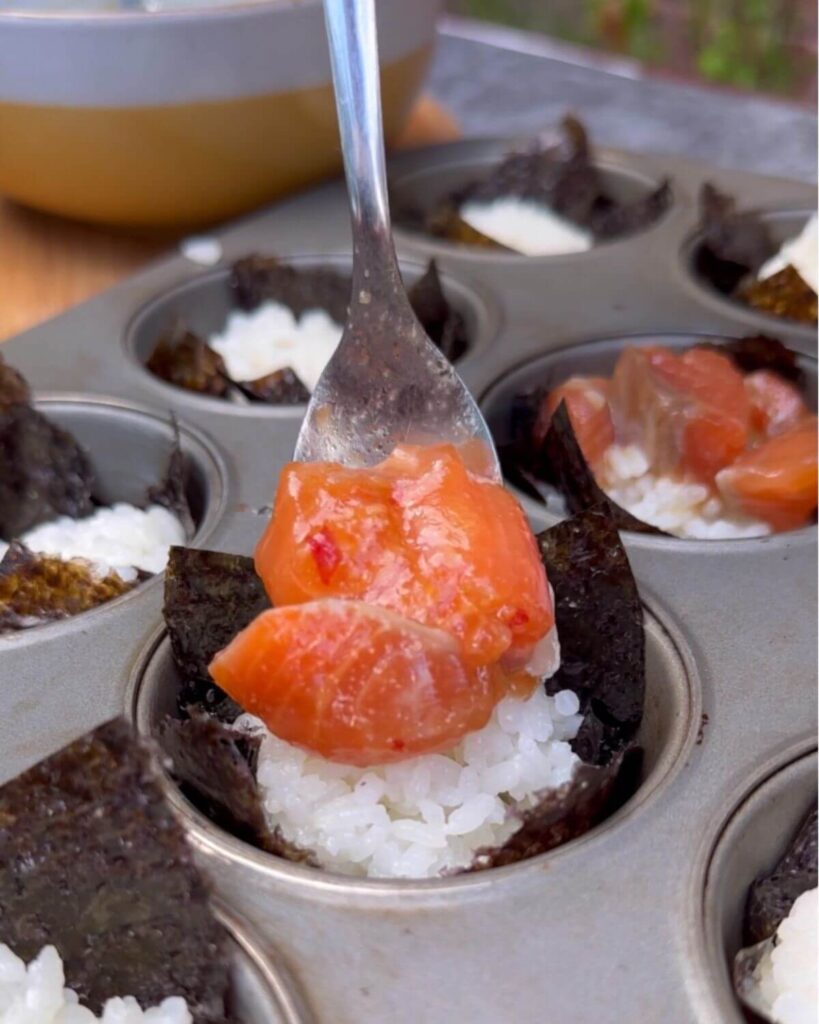 A close-up image of raw marinated salmon pieces being carefully placed on top of sushi rice-filled nori cups, arranged in a muffin tray, ready to be baked.