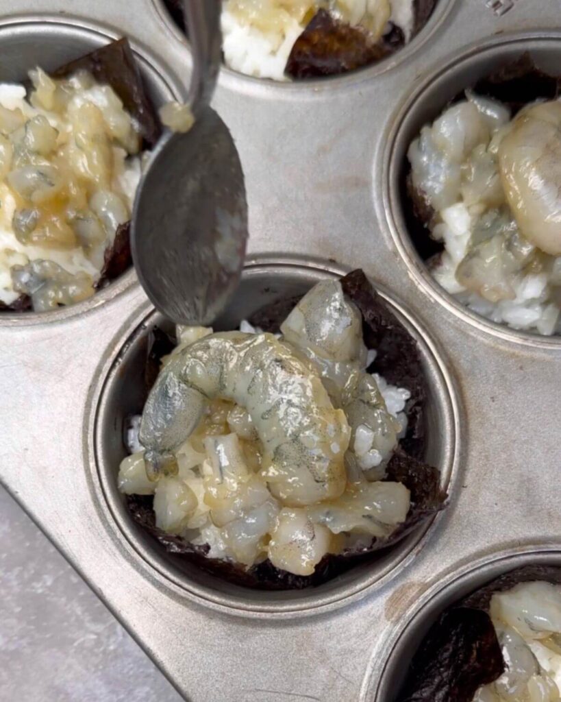 A close-up image showing raw marinated shrimp pieces being placed on top of the sushi rice in the nori cups, ready for baking. Some sushi cups have whole shrimp while others have smaller pieces, neatly arranged in a muffin tin.