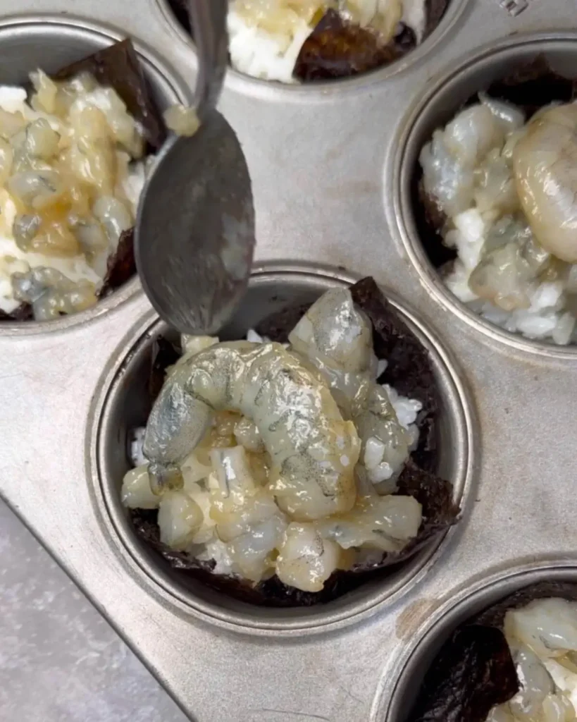 Close-up image of raw marinated shrimp being placed on sushi rice in nori cups, ready for baking. The muffin tin features cups with whole shrimp and others with smaller shrimp pieces, all neatly arranged.
