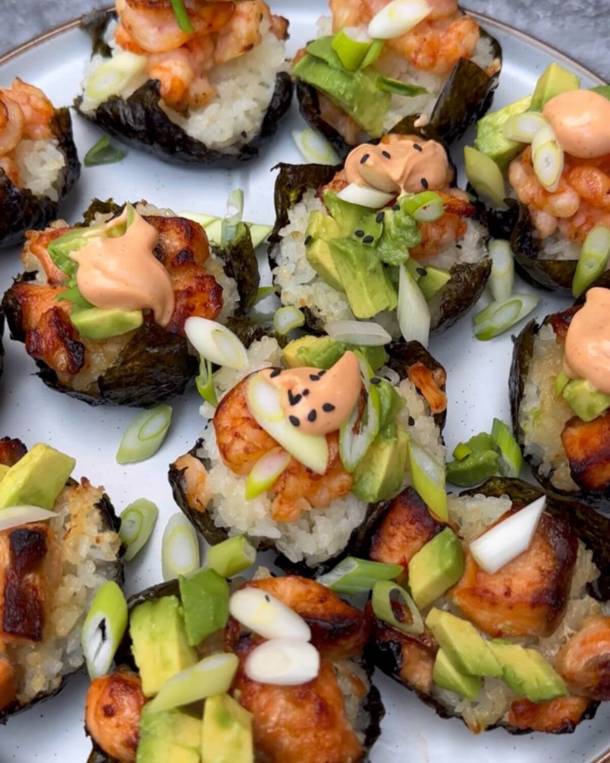 A photo showing freshly baked sushi cups, garnished with sesame seeds, sliced green onions, avocado cubes, and a drizzle of sriracha mayo, just taken out of the oven. The edges of the nori are slightly crispy, and the salmon and prawns are perfectly cooked.