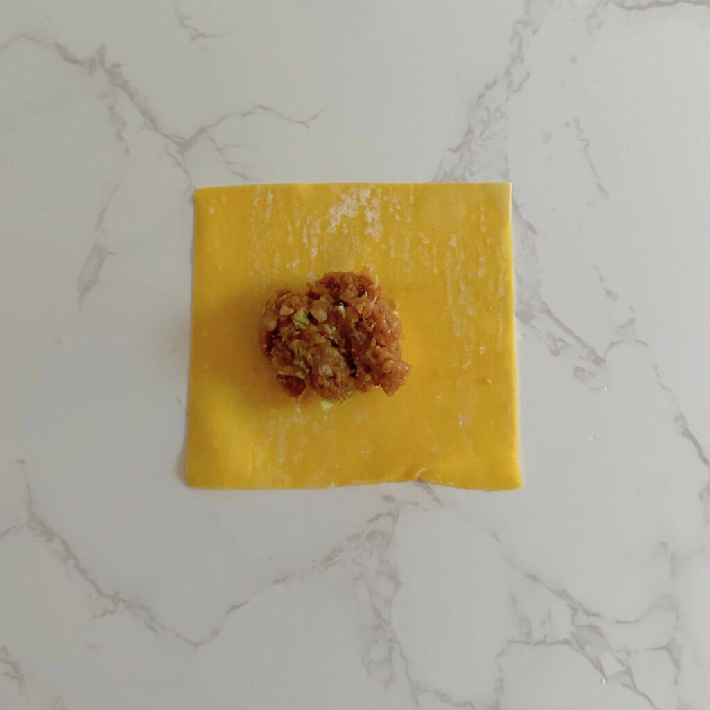 A square wonton pastry on a surface with a small amount of turkey filling in the center, ready to be folded.