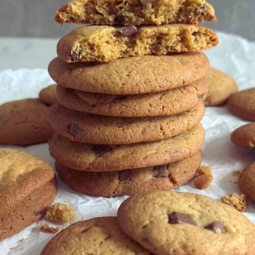 A stack of chewy chocolate chip cookies forming a tower, with one cookie split in half to reveal its soft, gooey center, surrounded by more cookies scattered around the tower.