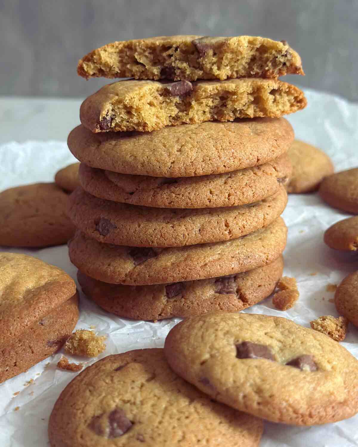 A stack of chewy chocolate chip cookies forming a tower, with one cookie split in half to reveal its soft, gooey center, surrounded by more cookies scattered around the tower.