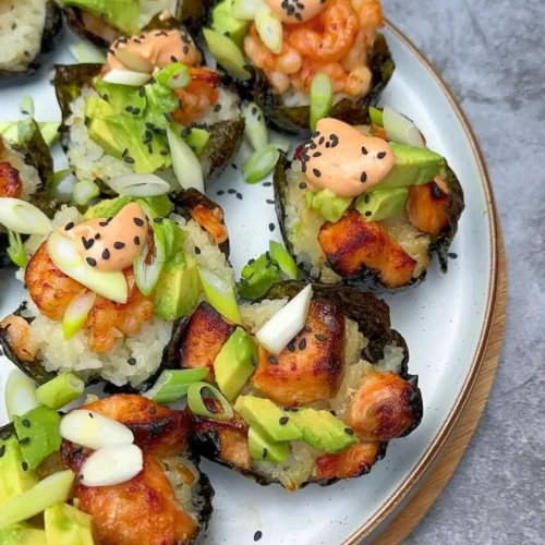 A close-up of baked salmon sushi cups, featuring crispy rice cups filled with tender baked salmon, topped with a mix of shrimp and salmon pieces. Drizzled with sriracha mayo, garnished with black sesame seeds, avocado cubes, and thinly sliced green onions, the sushi cups are beautifully arranged on a platter.